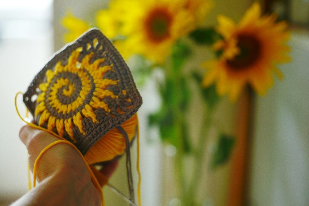 Hand holding a crocheted yellow sunflower motif with sunflowers in the background