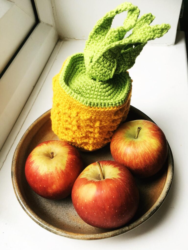 Yellow crocheted pineapple pencil case with green leaves beside apples